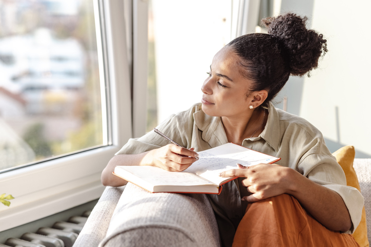 A woman sits by a sunlit window, writing in a notebook while looking outside thoughtfully.